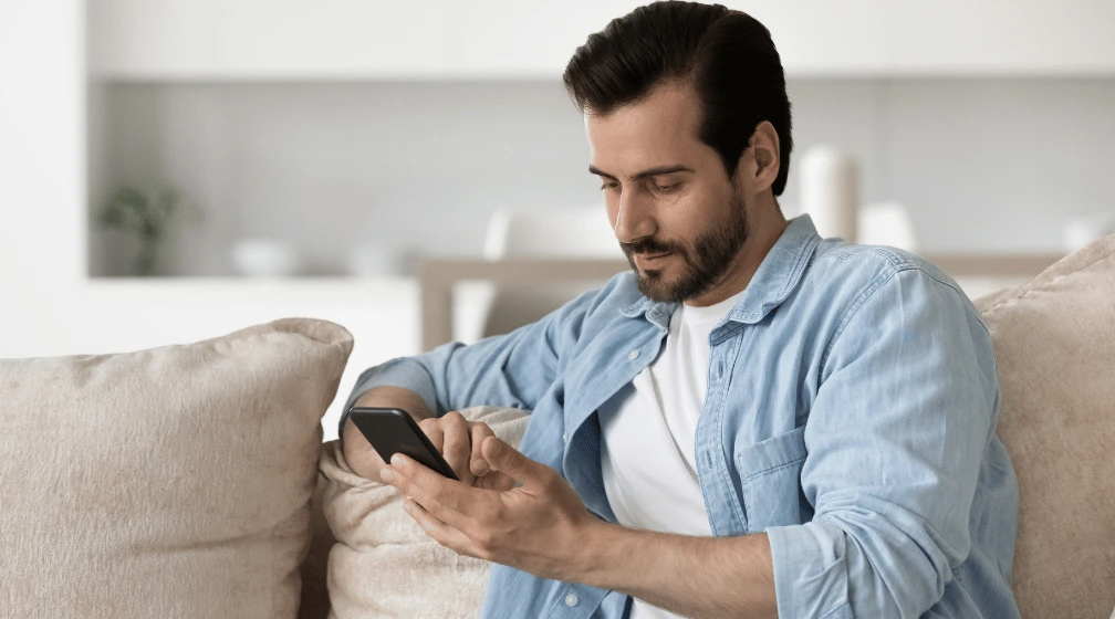 Imagem de homem sentado no sofá, olhando o celular, para ilustrar o artigo sobre "se não pagar multa de trânsito, o que acontece"