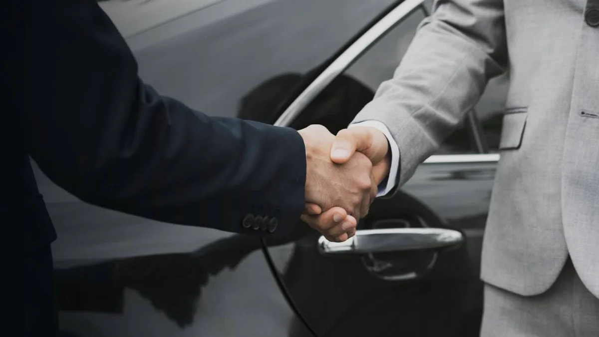 Aperto de mão entre duas pessoas vestindo ternos, com carro preto ao fundo. Conteúdo sobre transferência de veículos