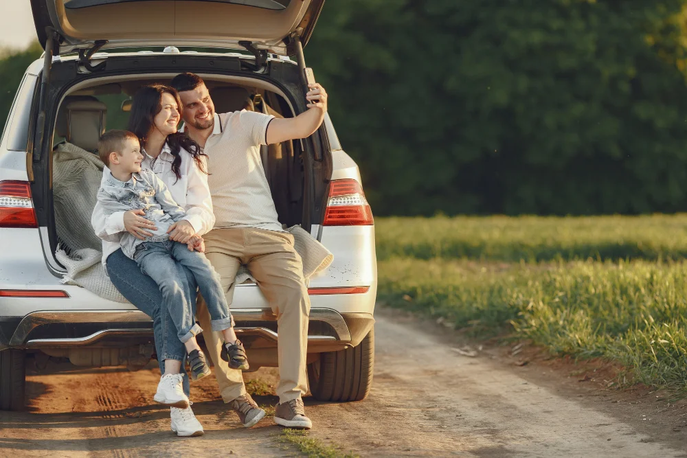 Família feliz sentada no porta-malas de um carro branco em estrada rural, tirando selfie ao pôr do sol. Conteúdo sobre como planejar uma viagem de carro